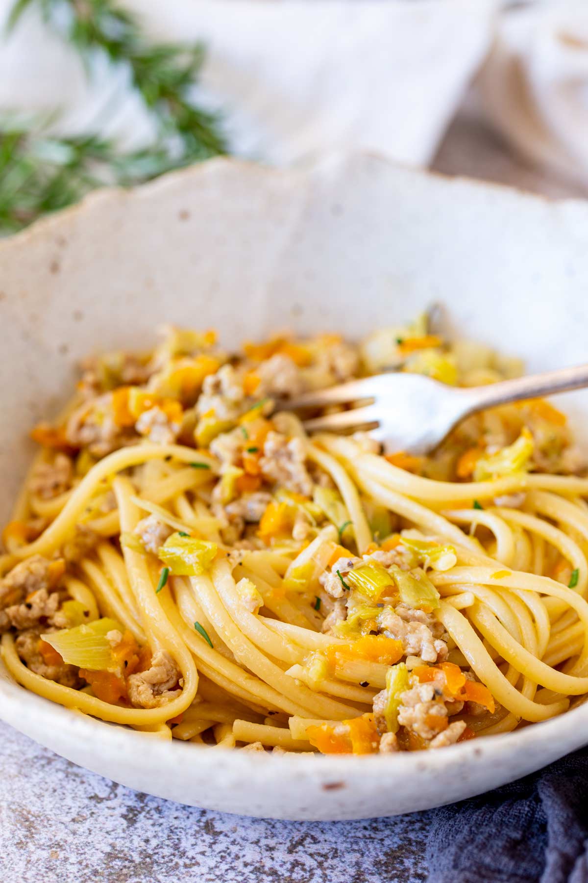A rustic ceramic bowl filled with pasta in a leek and pork sauce, with a fork resting at the back and a textured beige napkin.