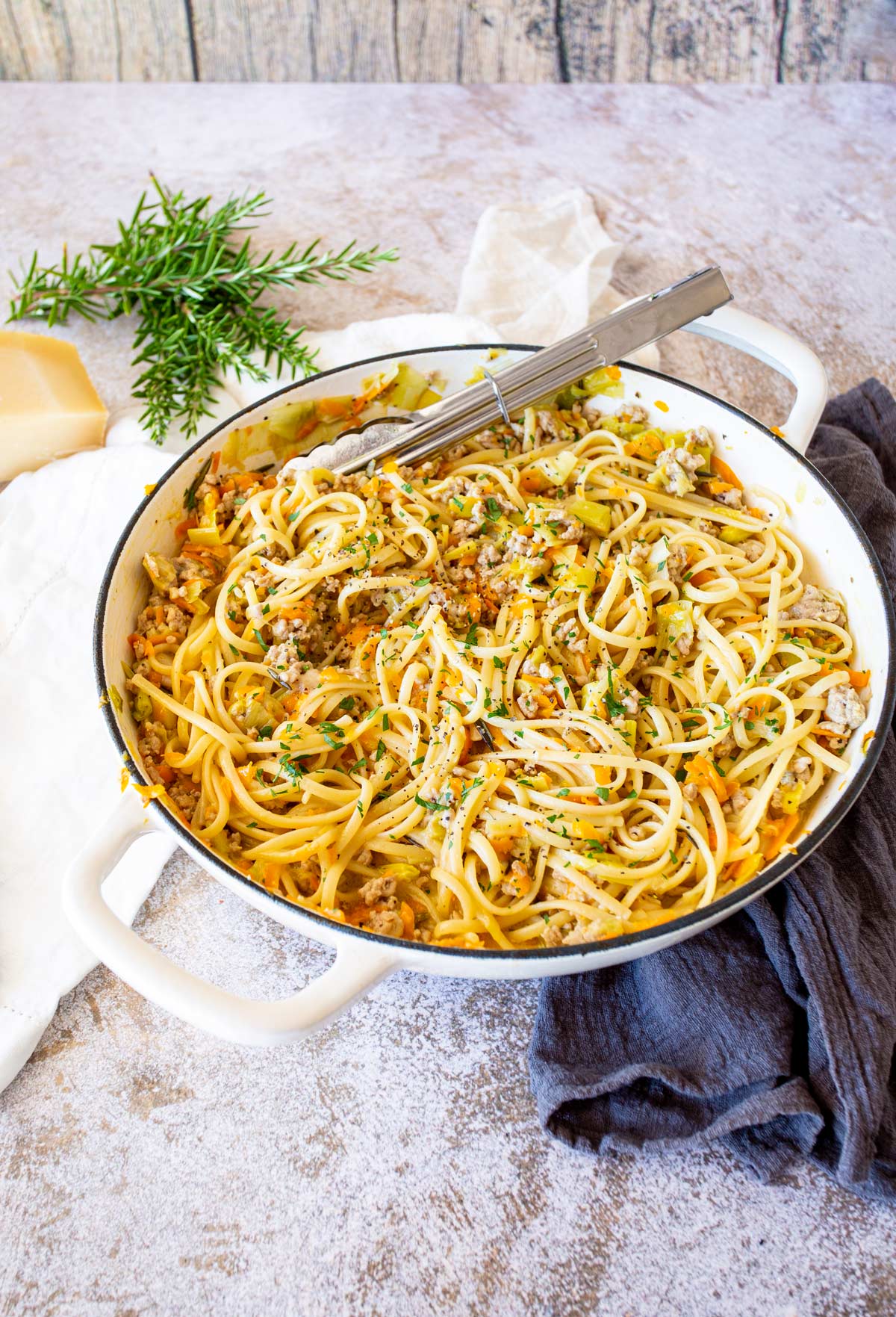 A full table view with a white cast iron pan filled with linguine and pork, surrounded by a rustic setting with linens and fresh rosemary.
