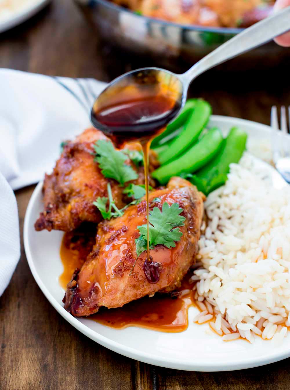 a spoon drizzling a sticky golden sauce over a cooked chicken thigh on a plate with rice