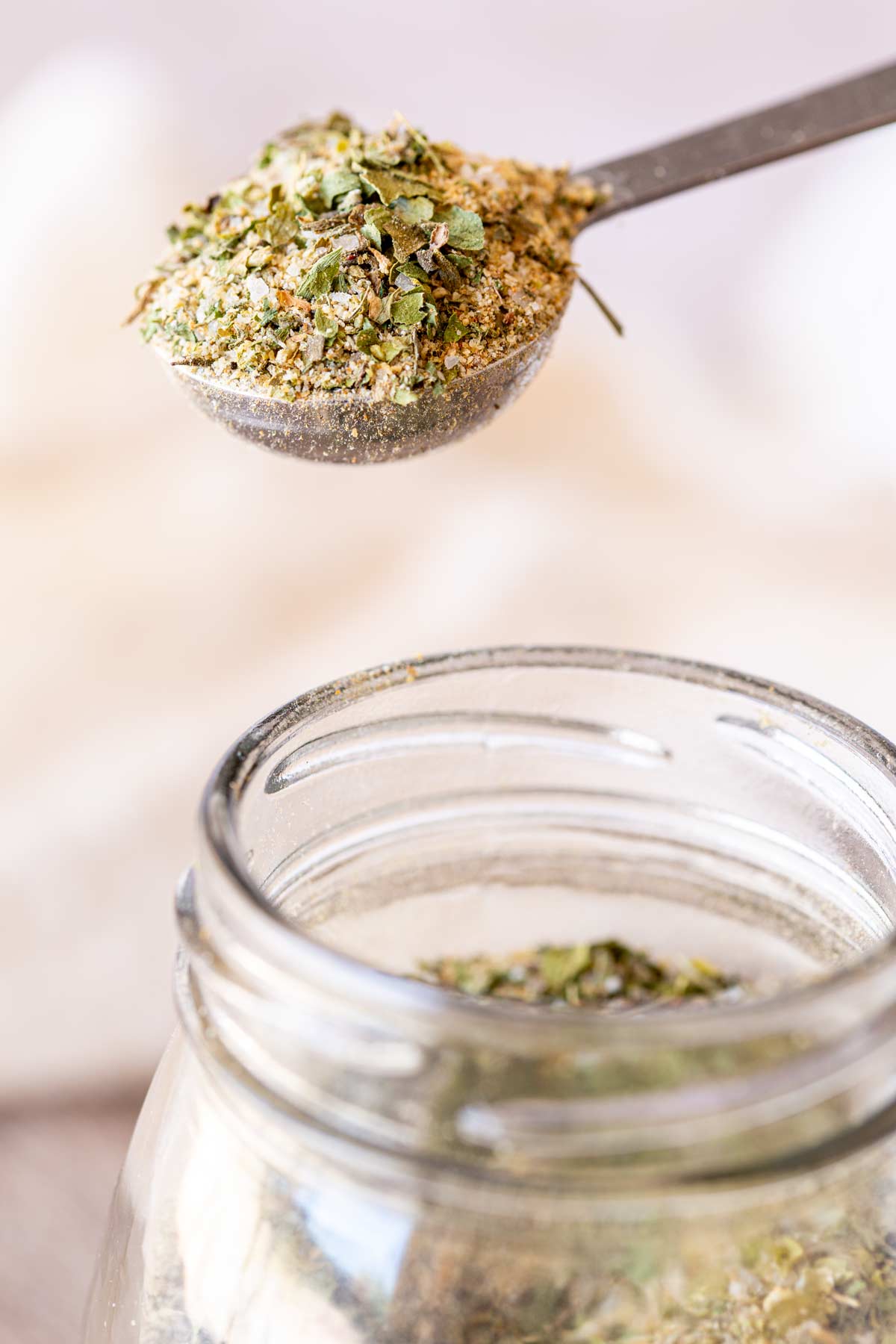 A measuring spoon heaped with garlic and herb seasoning, held above a glass jar filled with the spice blend.