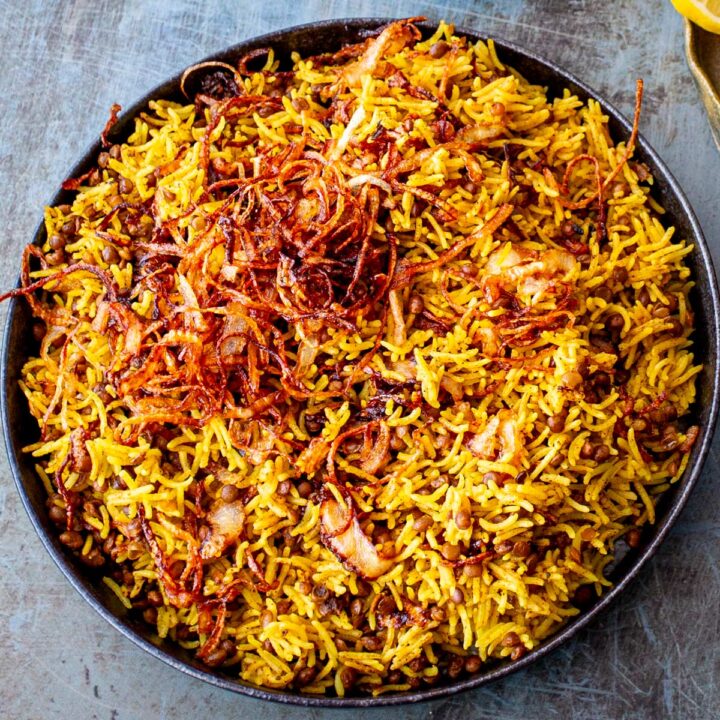 A bowl of Mejadra, with spiced lentils and rice, served in a dark grey bowl on a blue/silver-grey table.