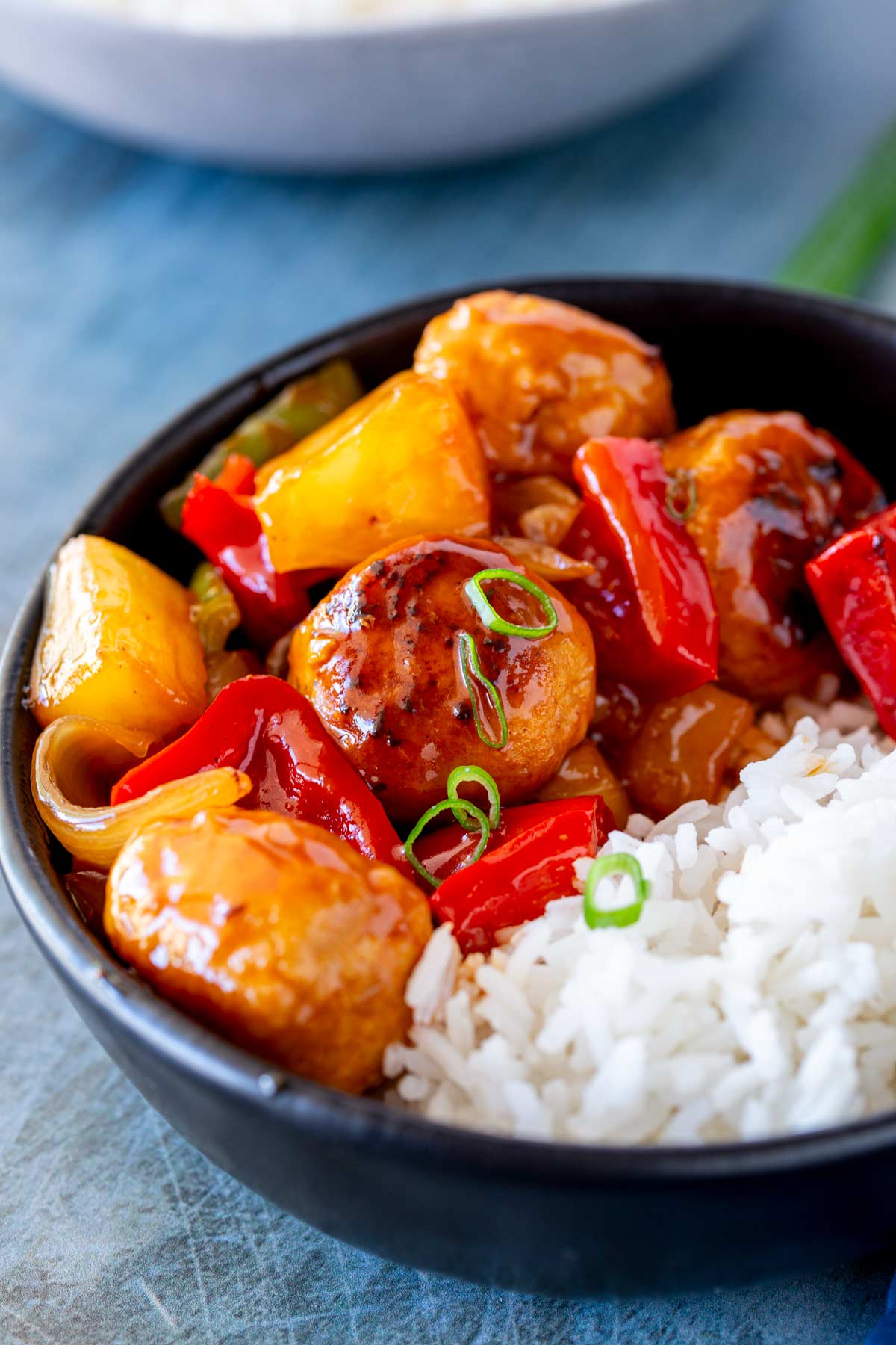 A bowl of sweet and sour chicken meatballs coated in a glossy sauce, served with white rice and garnished with green onions.