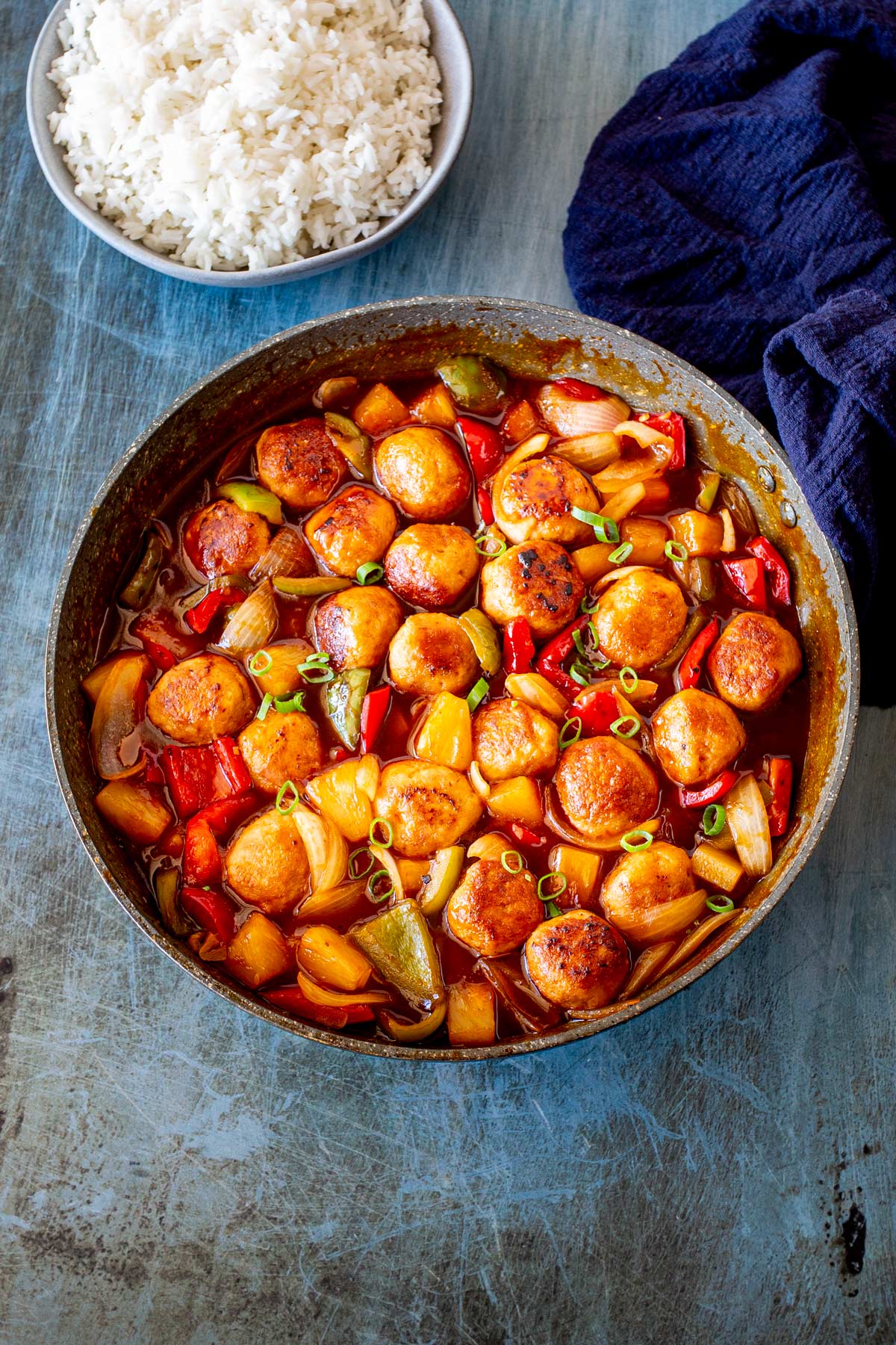 A large pan filled with meatballs coated in a glossy sauce, mixed with red and green bell peppers. with a blue napkin and table