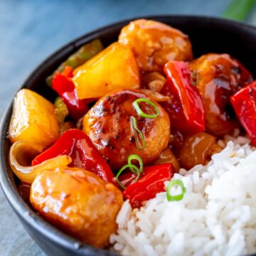 A close-up of chicken meatballs in a black bowl, with a sticky sauce bell peppers and pineapple, with white rice and sprinkled with chopped green onions.