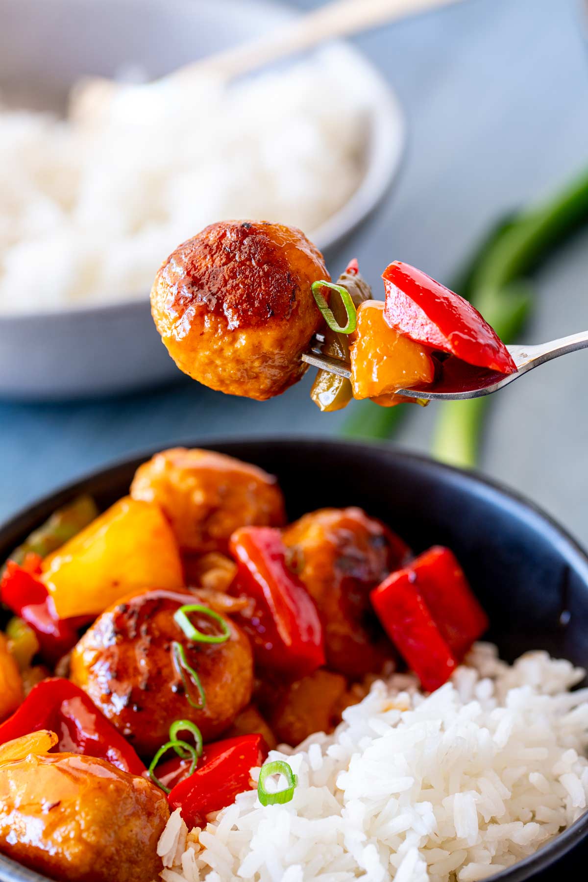 A single chicken meatball and chunks of bell peppers held by a fork over a bowl of sweet and sour with rice