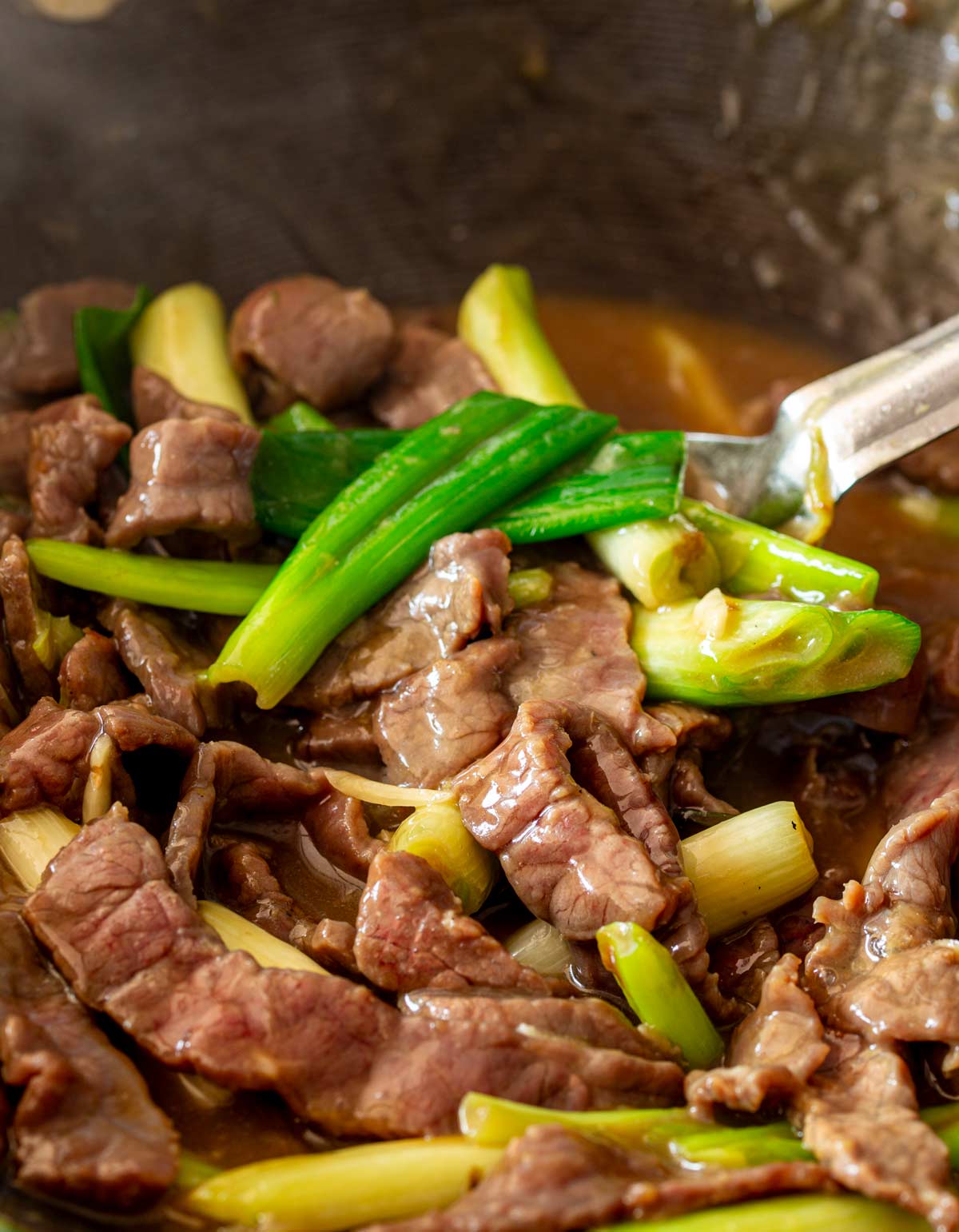 A metal serving spoon scooping up glossy strips of beef and vibrant spring onions coated in brown sauce