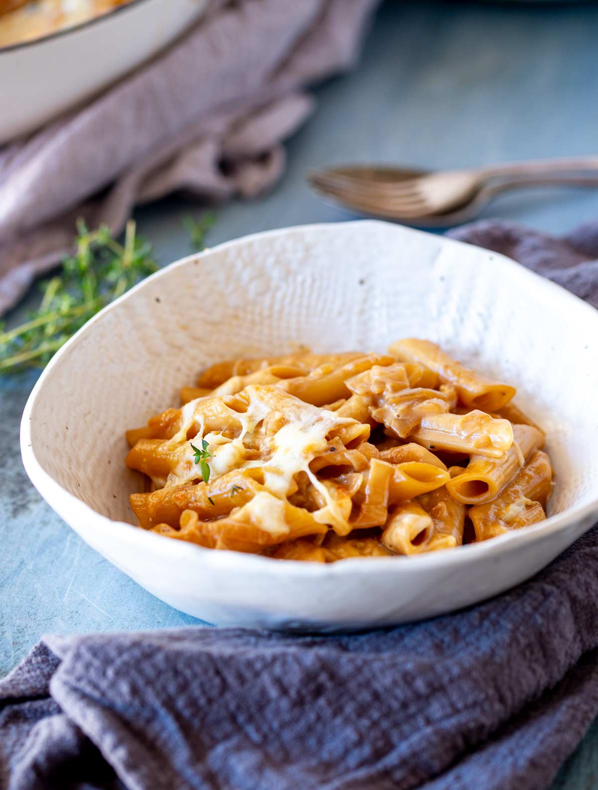Side-angle photo of a white bowl of pasta on a scratched blue metal surface, with a linen napkin and cutlery in frame