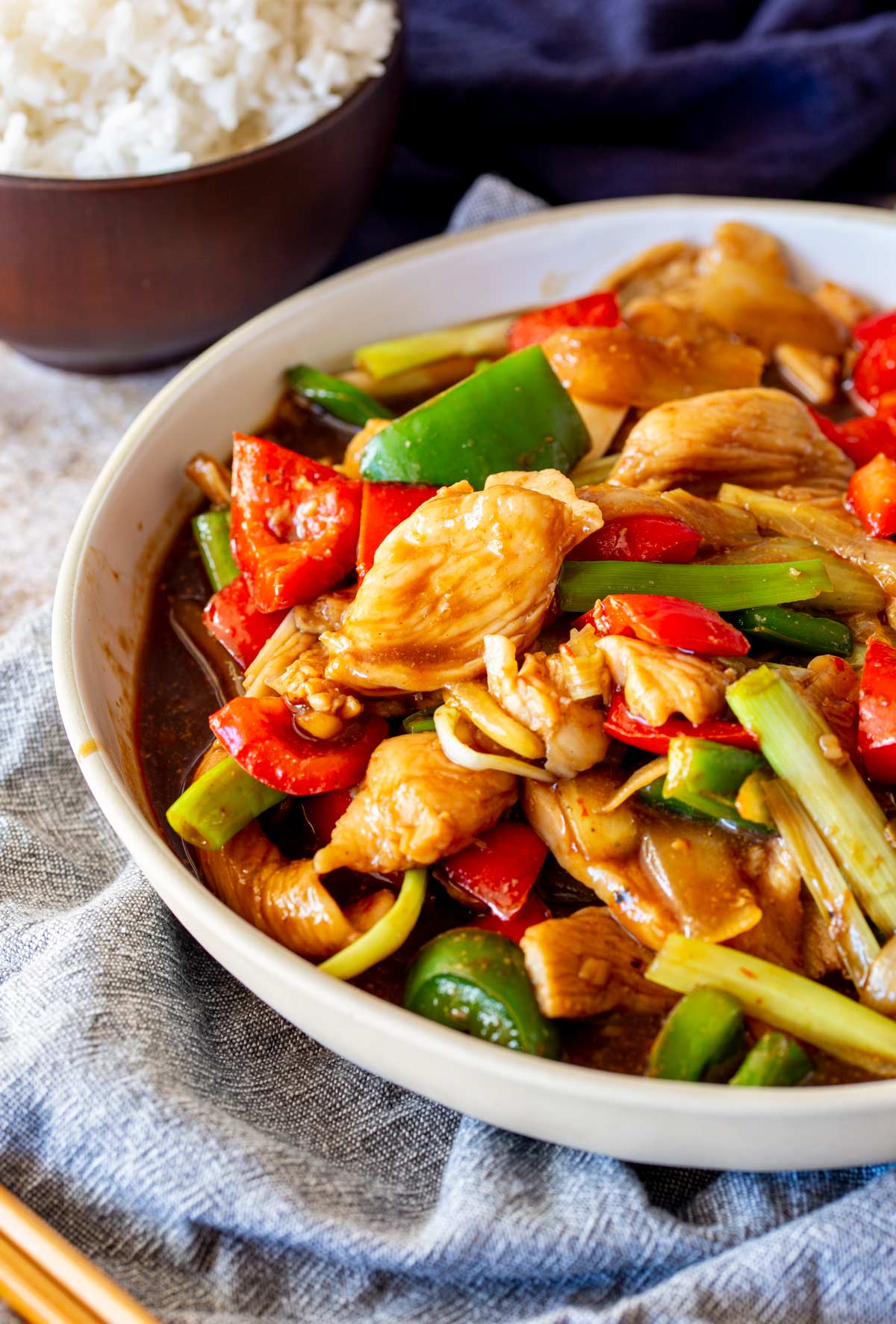 Close-up of glossy stir-fried chicken and vegetables in a beige bowl, with white rice and a blue napkin in the background