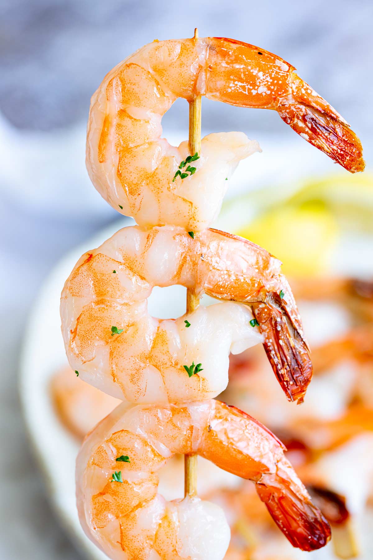 One shrimp skewer being held vertically, with three shrimp on the wooden stick and a plate of skewers blurred in the background