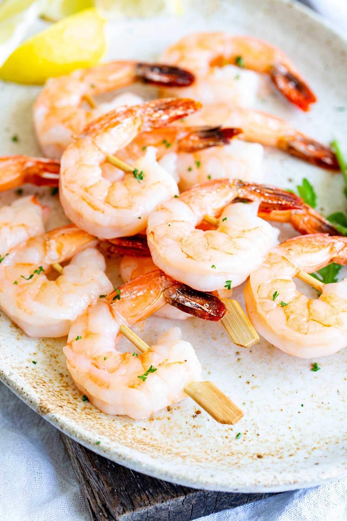Three shrimp on wooden skewers, plated on a rustic oval dish with salad leaves, lemon wedges, and a sprinkle of parsley