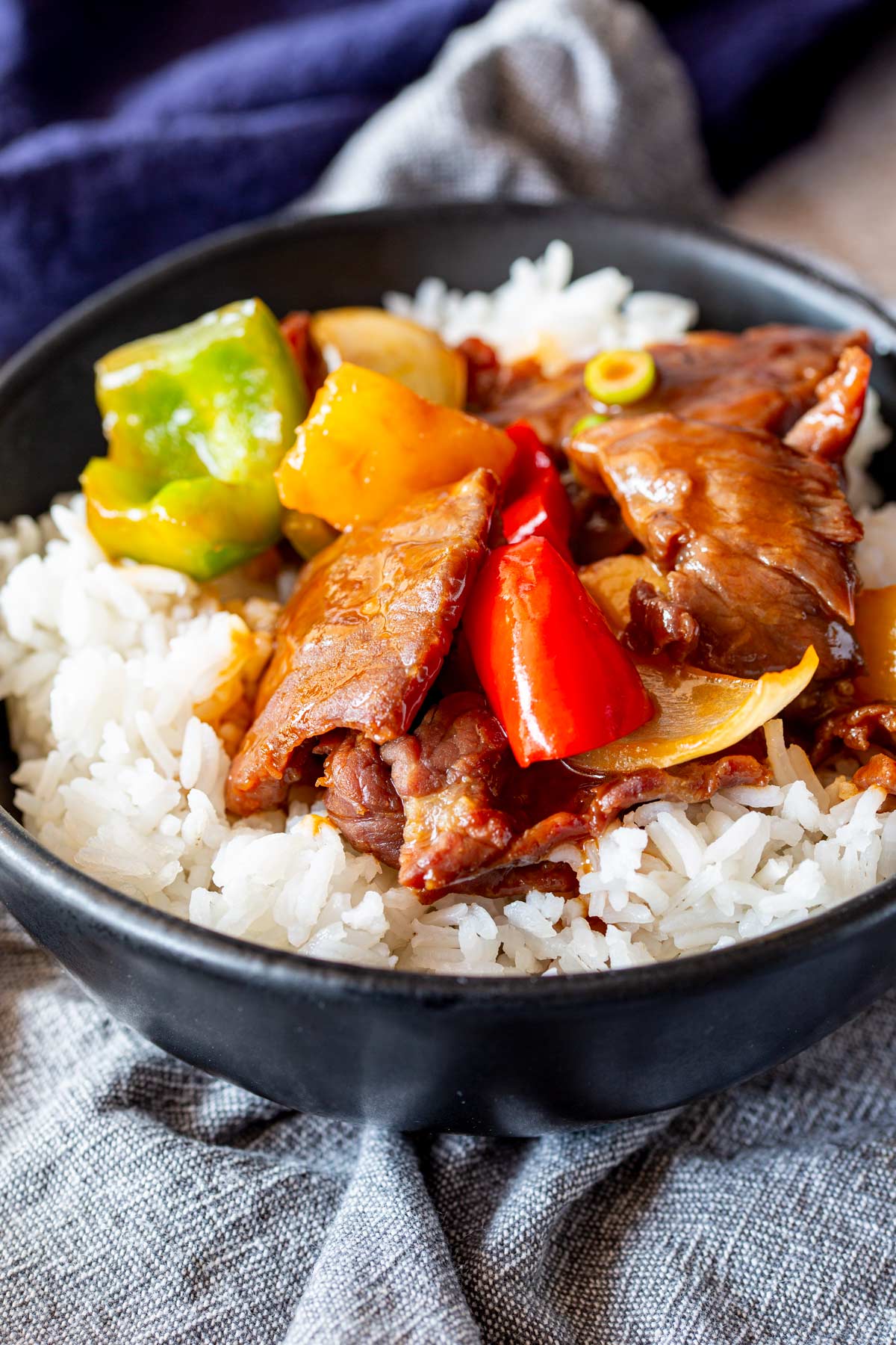 Glossy strips of beef stir fry served over rice in a black bowl, styled with a grey linen napkin
