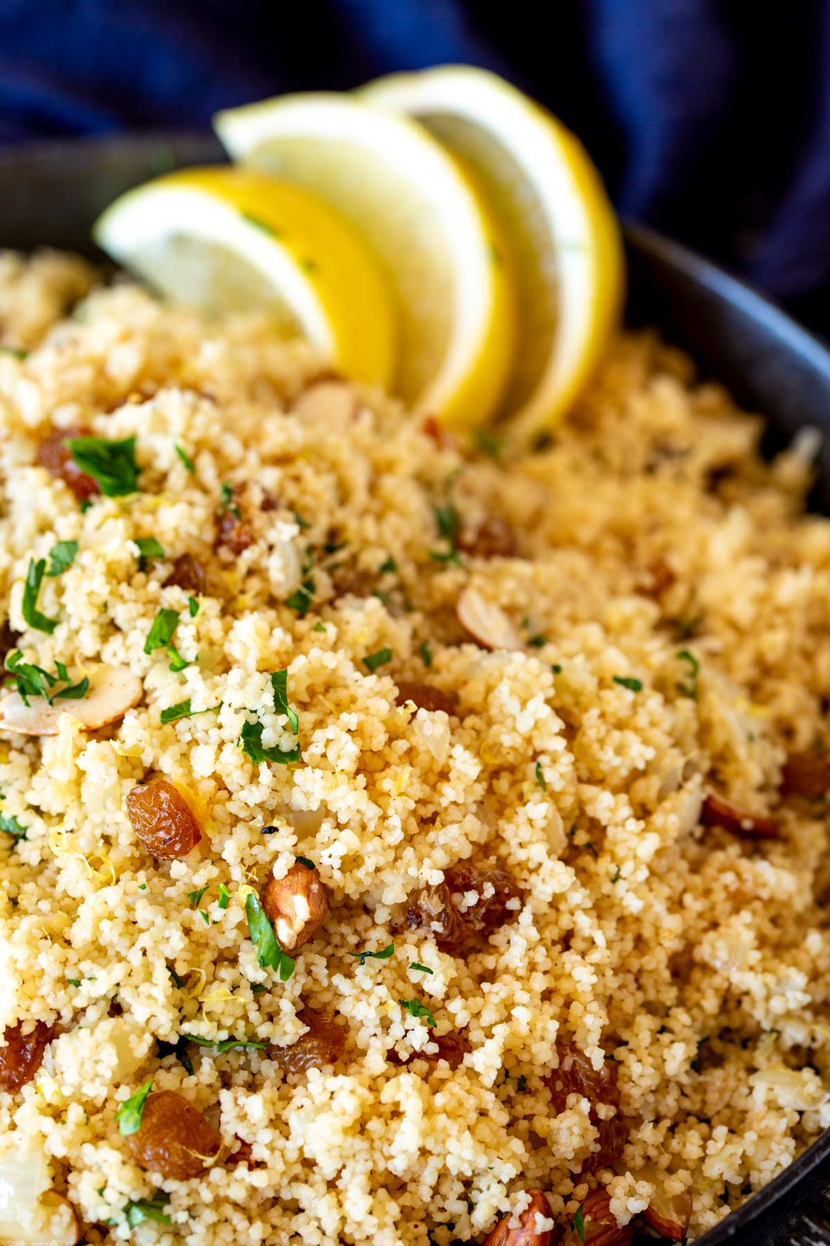 close up on Cooked couscous with lemon zest, dried fruit, and chopped herbs. Blurred lemon slices in the background