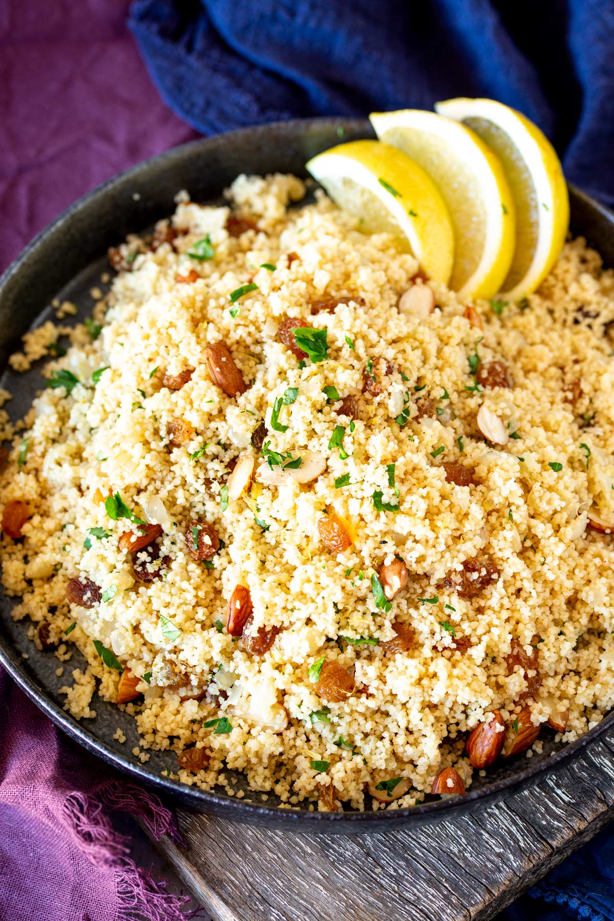 overhead of couscous in a dark shallow bowl with raisins and almonds mixed through it, lemon slices and a purple napkin