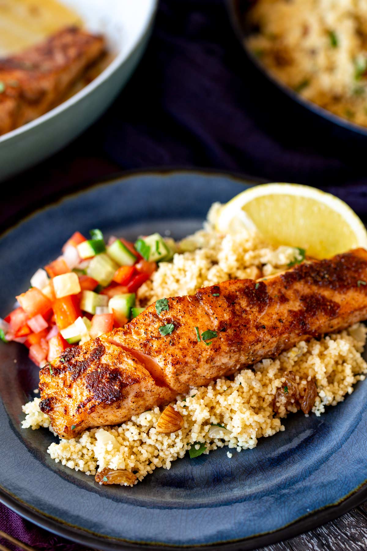 a cooked fillet of salmon on a bed of couscous on a blue plate with a chopped salad and lemon wedge