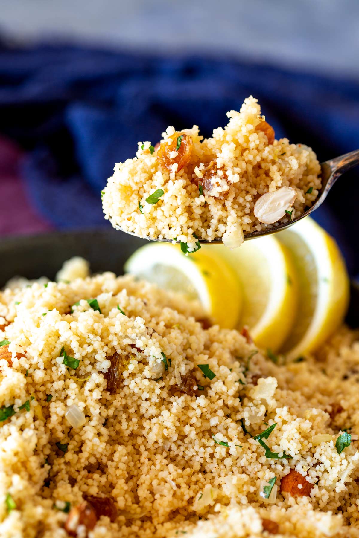 A spoon lifting up a serving of cooked couscous dotted with dried fruit and nuts