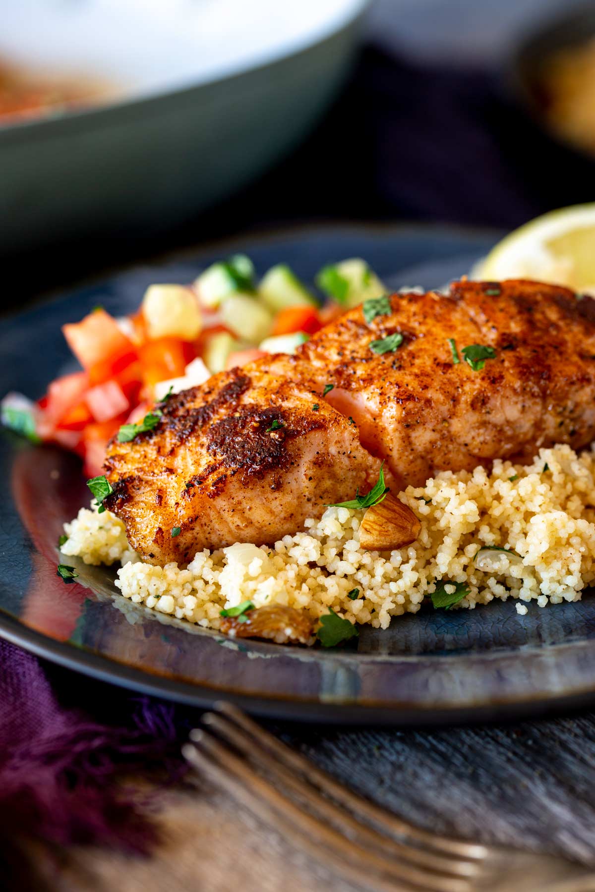 Small blue plate holding salmon, couscous with nuts and fruit, a tomato salad, and lemon wedges on a rustic wooden table