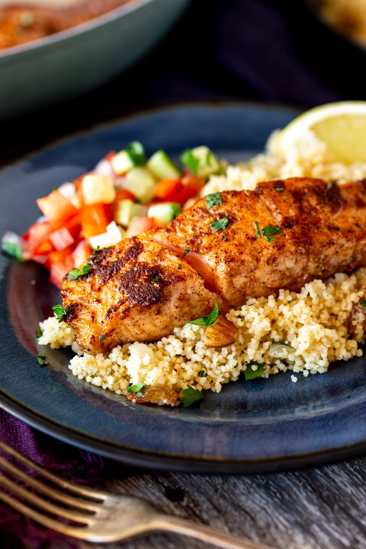 Single salmon fillet on a blue plate with herbed couscous, chopped salad, lemon wedges, and a rustic wooden table set with a purple napkin