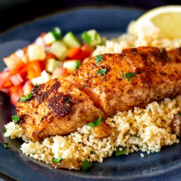 close up of a salmon fillet with lemon nut couscous, tomato salad, chopped herbs and a wedge of lemon