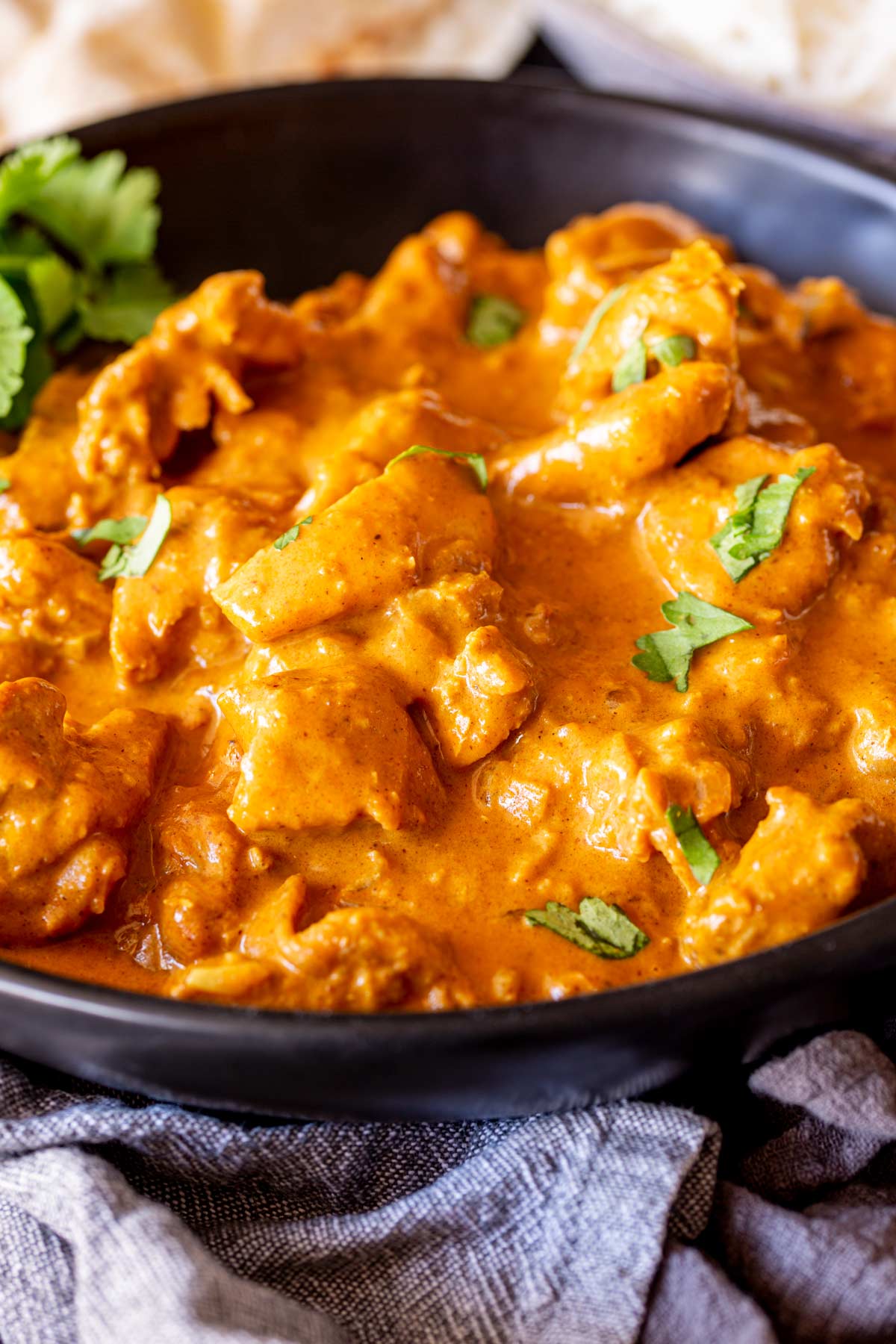 A black bowl of creamy Chicken Korma, rice and bread sit just out of focus, with grey linen