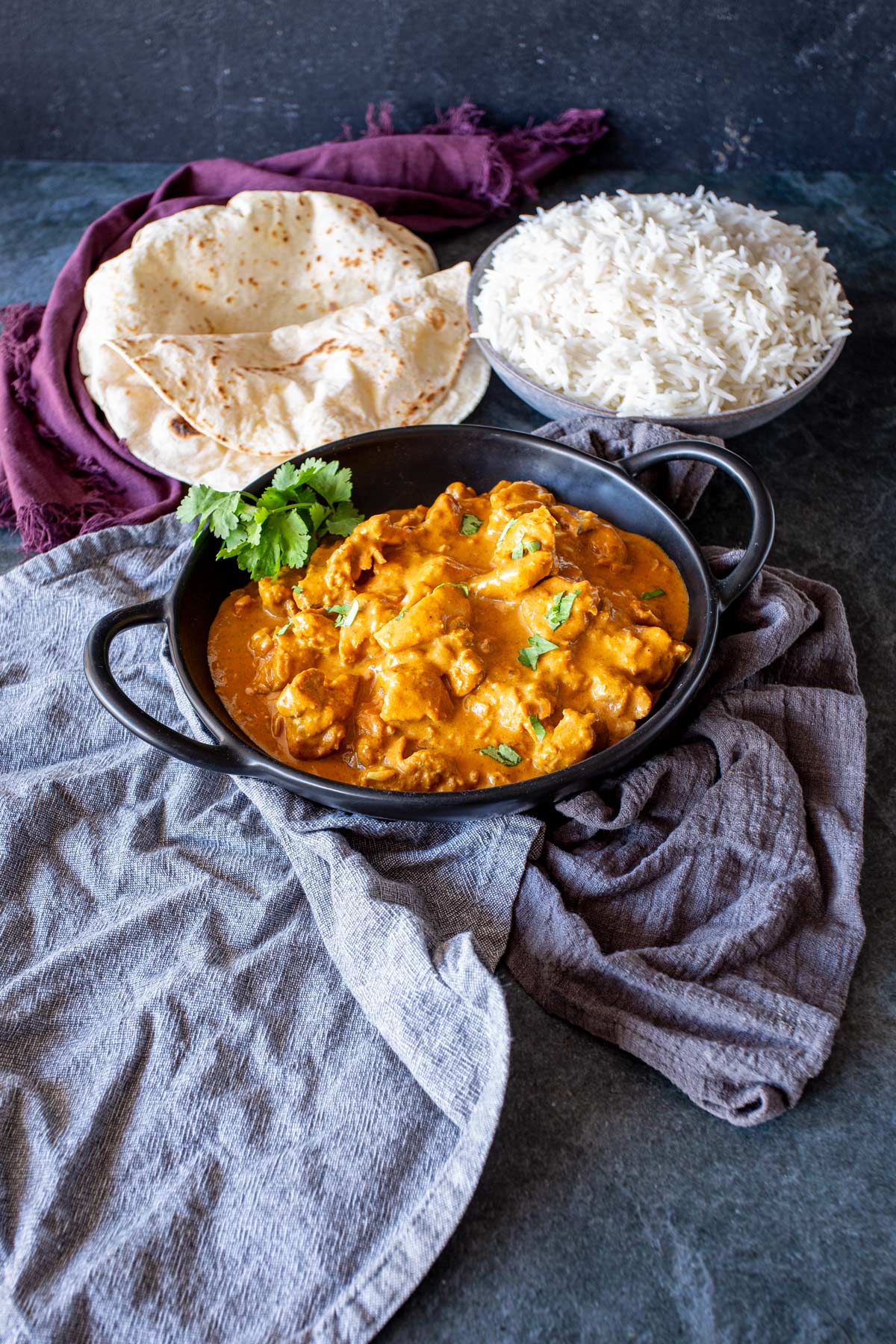 Black bowl of Chicken Korma served with rice and roti, on a dark countertop with soft grey and purple napkins