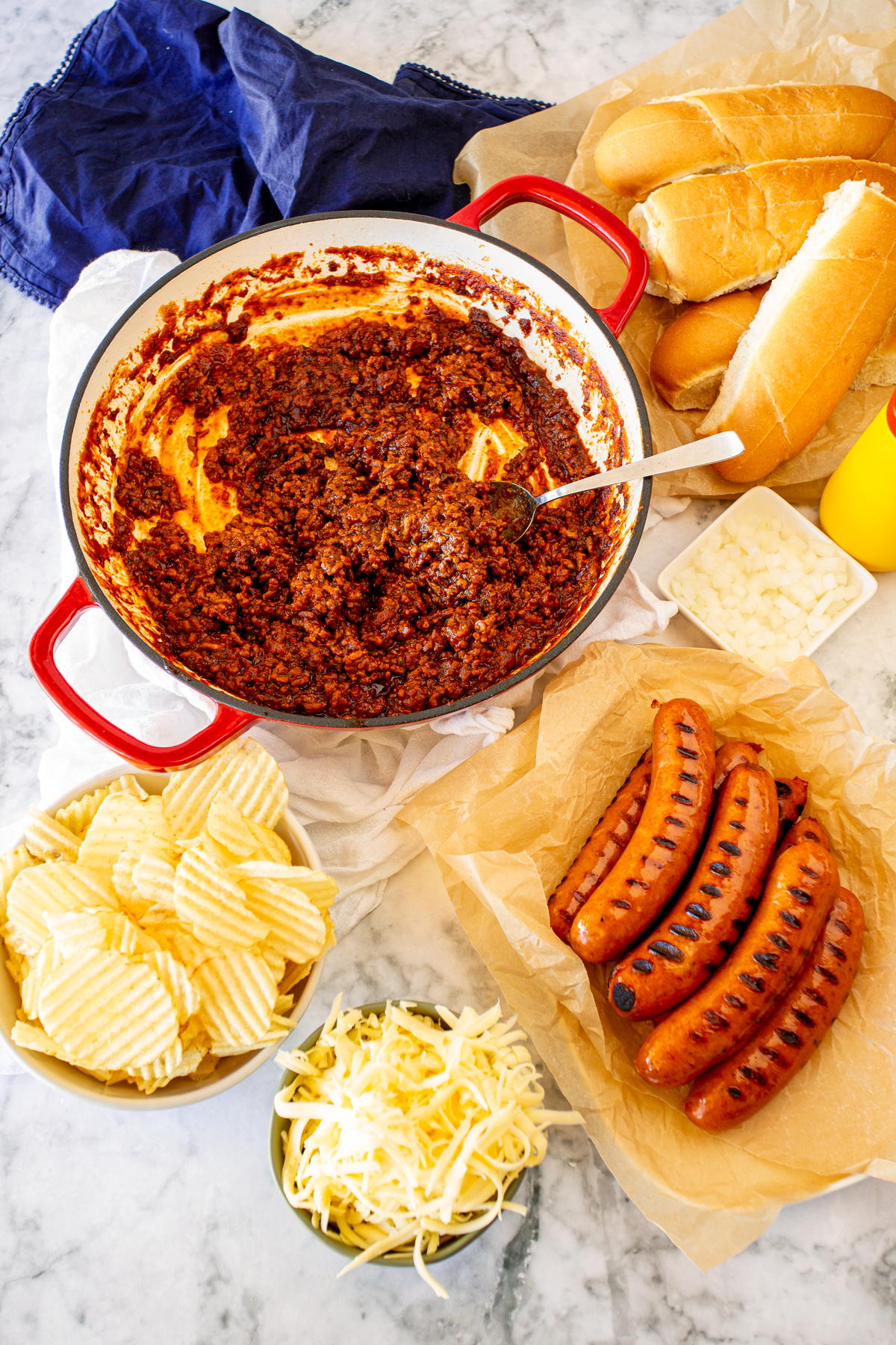 A table laid out with chili sauce, hot dogs, buns, cheese, onions, and chips