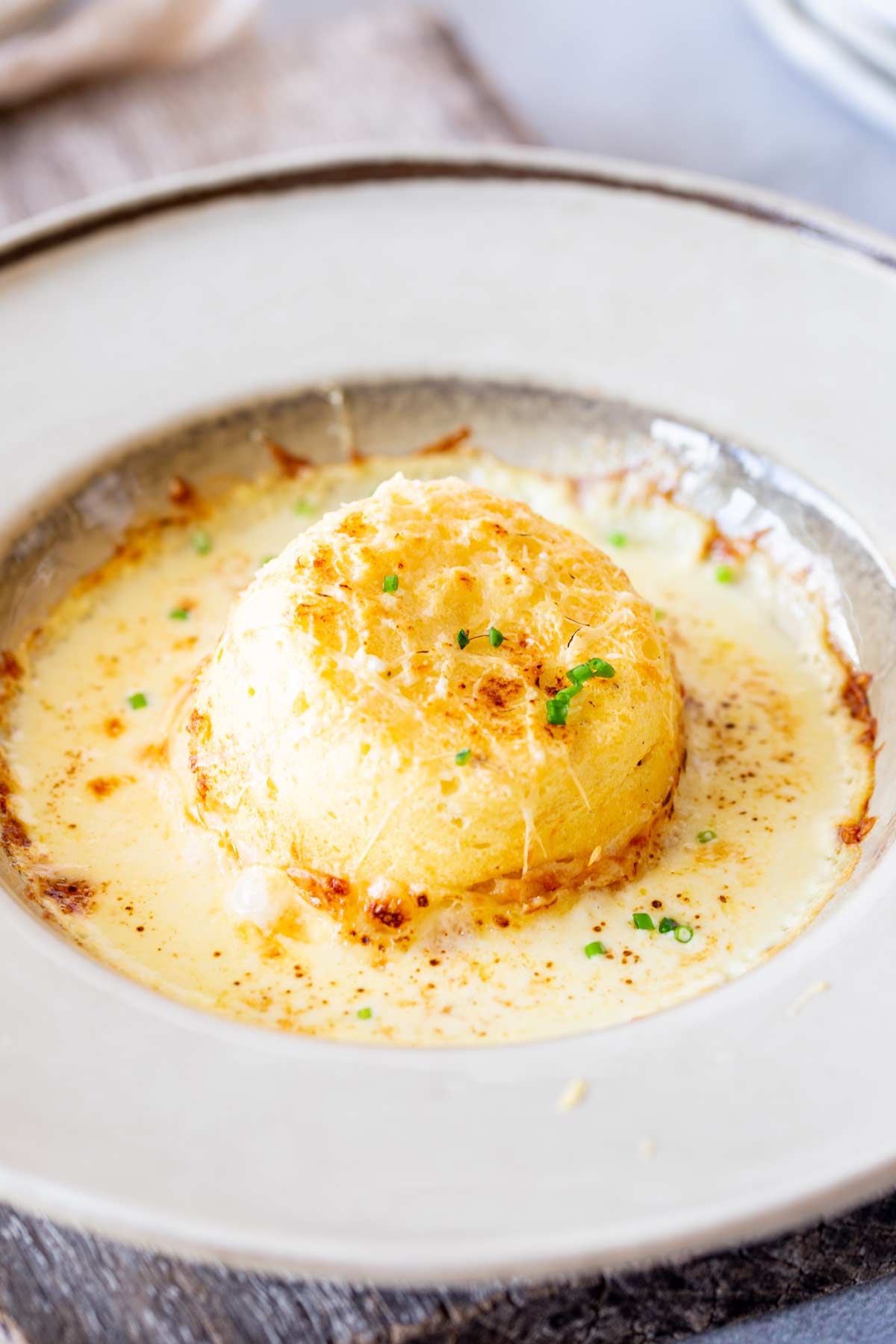 Single cheese soufflé in beige dish with a wooden board underneath.