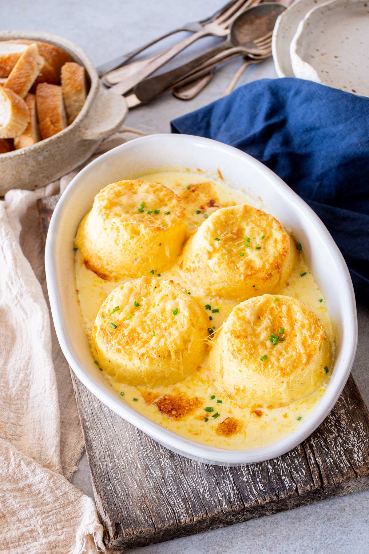 Golden twice baked cheese soufflés in a white rustic baking dish on a grey-blue counter with cream linen and vintage cutlery