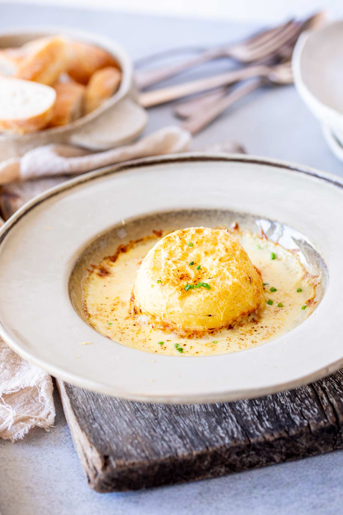Single cheese soufflé in beige dish with cream linens and bread in the background