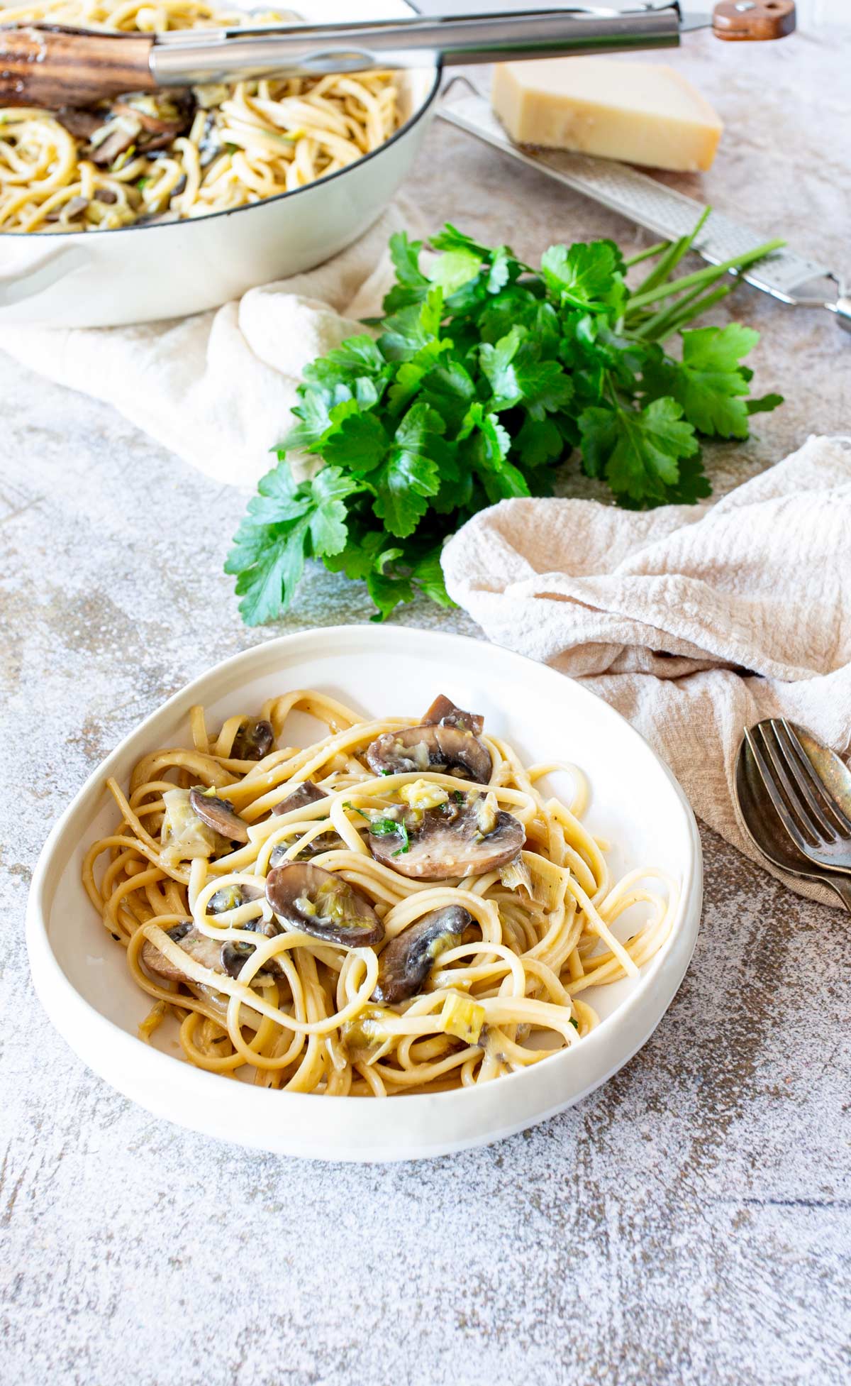 Cream bowl of pasta with mushrooms and leeks on a stone table with a bunch of herbs and a block of parmesan in the background
