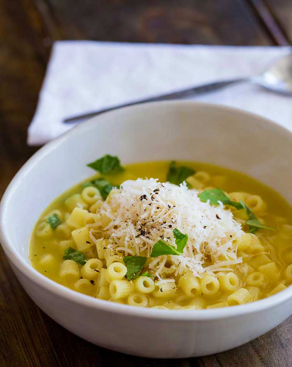 a white bowl of pasta in broth on a wooden table with a napkin and spoon