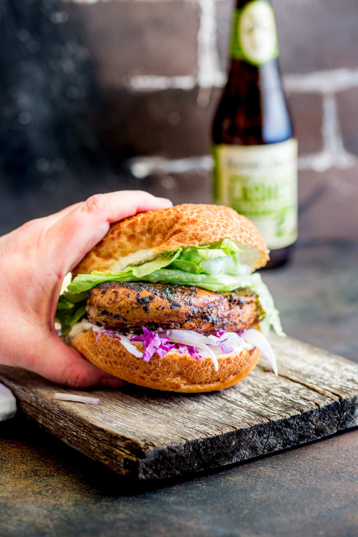 A hand holding a chicken burger with lettuce in a bun, sitting on a wooden board with a drink bottle behind