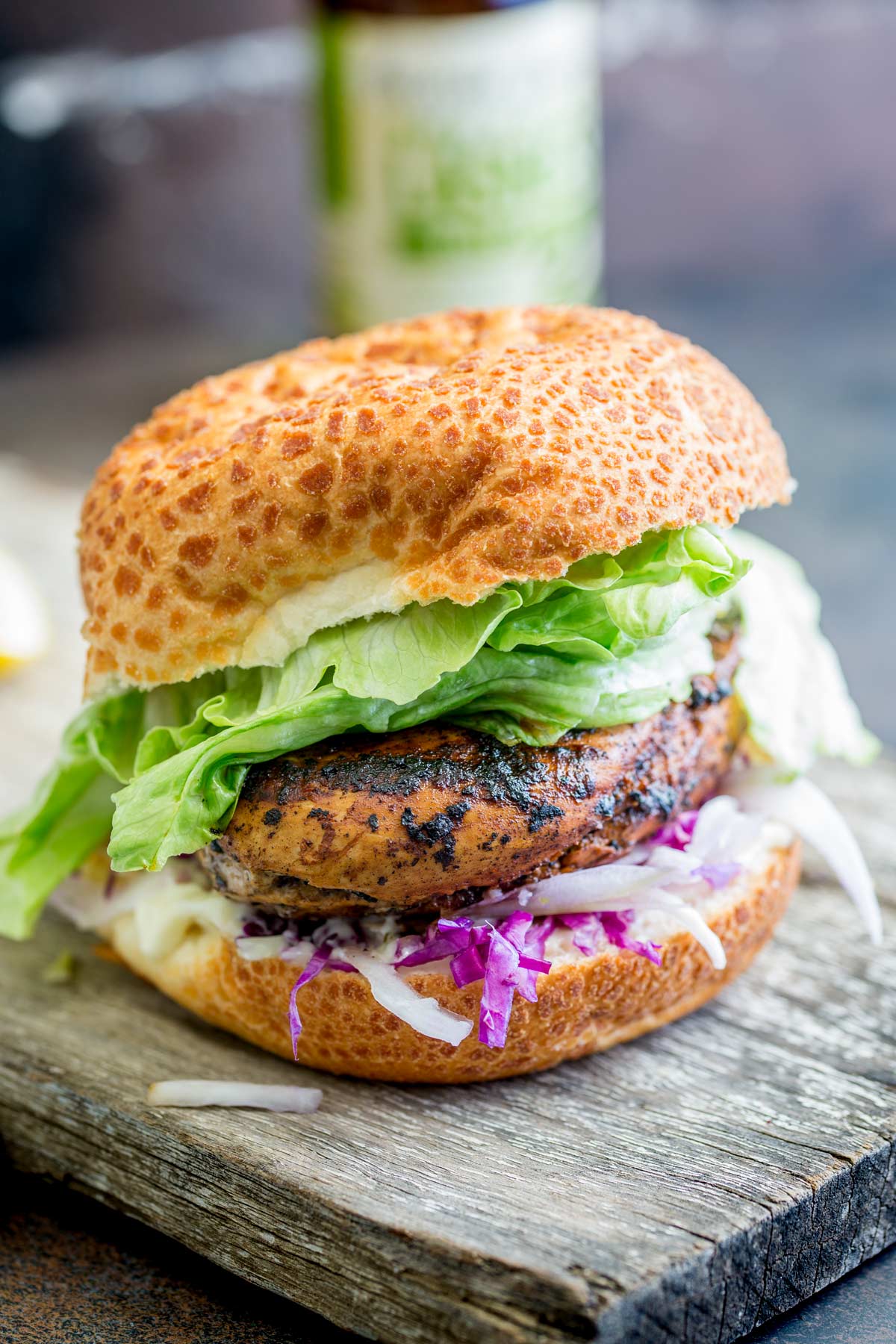 Chicken burger with lettuce in a bun on a wooden board, with a bottle of drink and blurred background elements