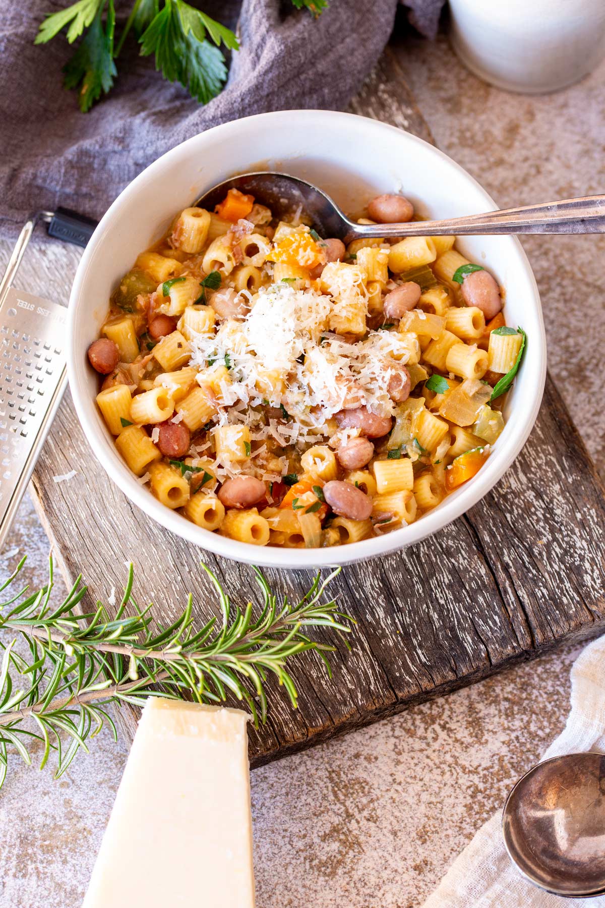 a white bowl of bean and pasta soup on a rustic board with herbs on it