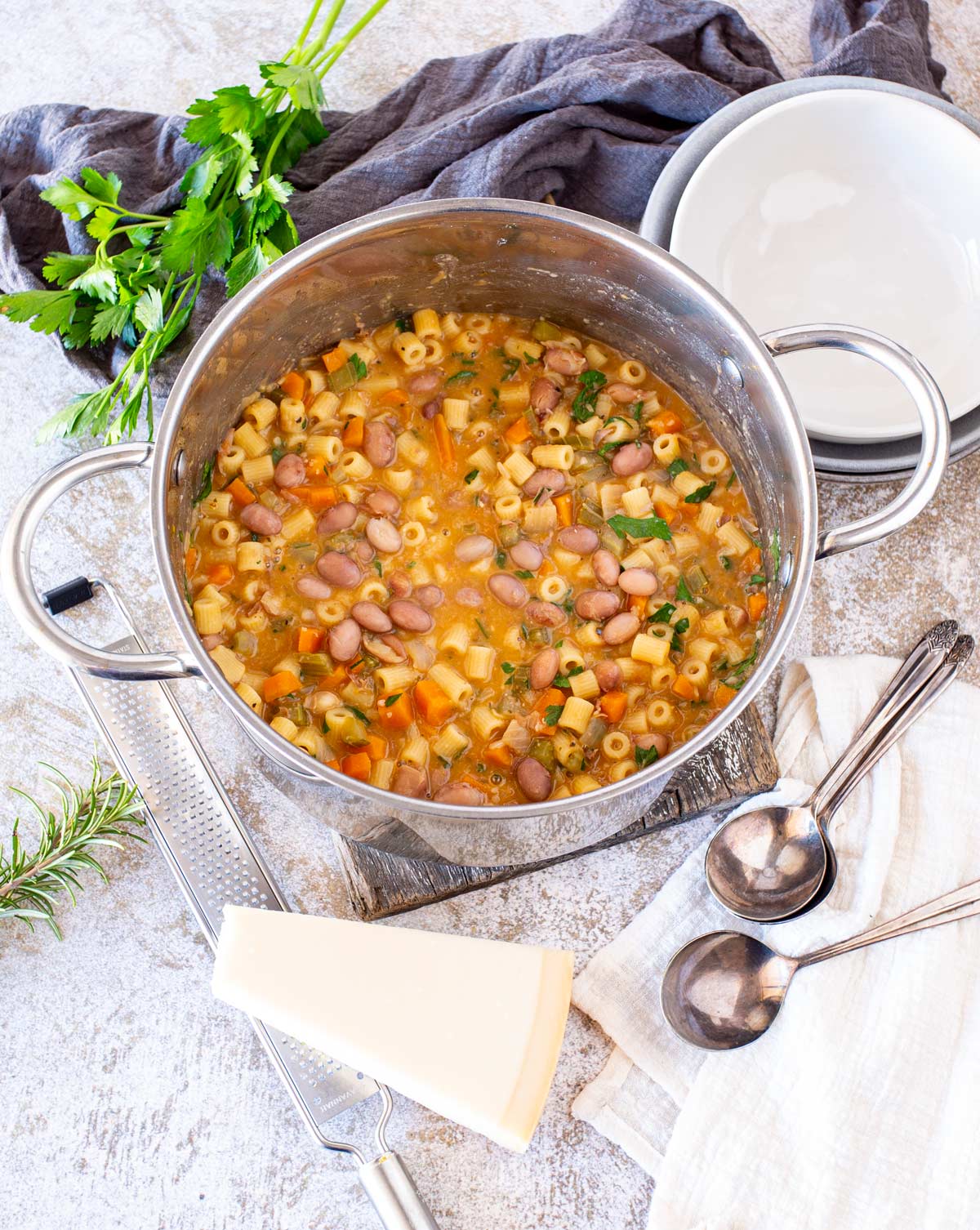 a pan of soup on a rustic board on a table with herbs, cheese, bowls and spoons