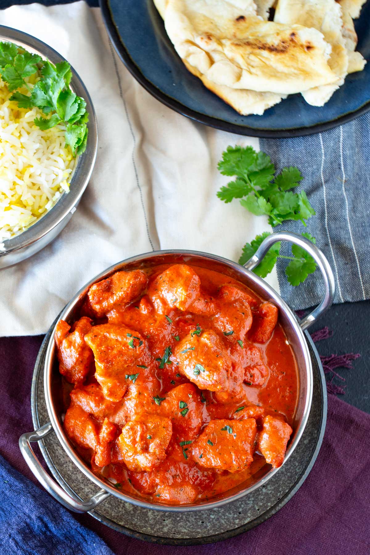 Overhead view of Chicken Chasni in a traditional metal curry dish surrounded by fluffy rice, torn naan bread, and colorful blue and purple napkins