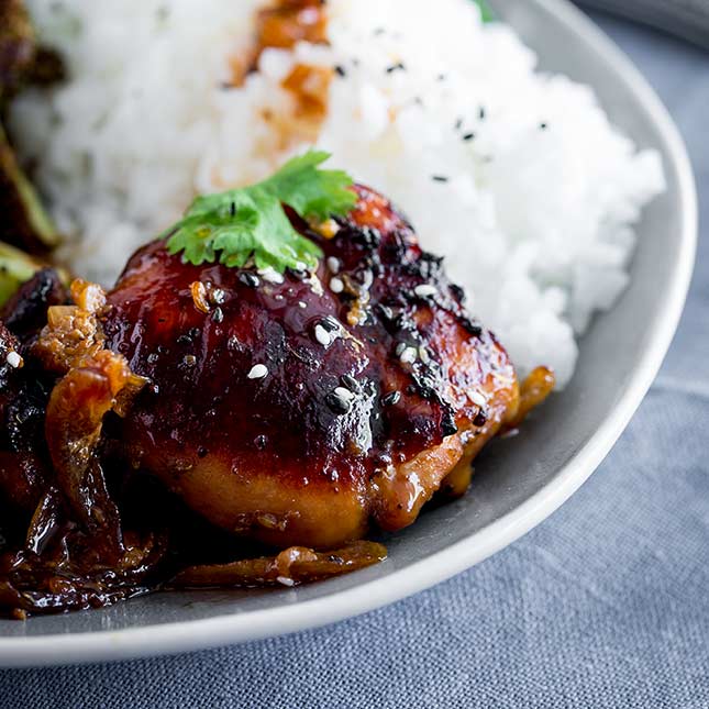 close up of a sticky chicken thigh on a grey plate with rice behind