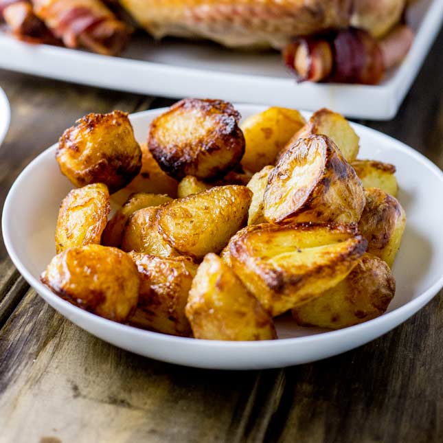 Crispy fluffy roast potatoes in a white bowl with a roast turkey behind